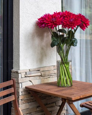 Burgonya kırmızısı yıldız çiçekleriyle dolu cam bir vazo terastaki ahşap bir masada oturup bahçenin güzelliğini arttırır..