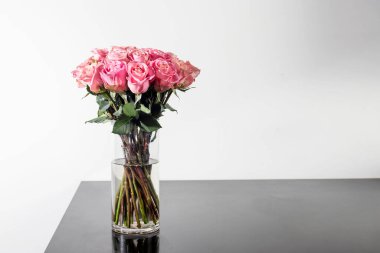 Siyah bir masanın üzerindeki cam vazoda beyaz bir arka planda duran pembe güllerden oluşan bir buket. Kopyalama Boşluğu