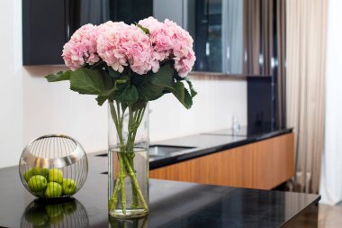 Cam silindirik vazoda pembe ortancalar ve mutfak tezgahının parlak yüzeyinde yeşil elmalar..