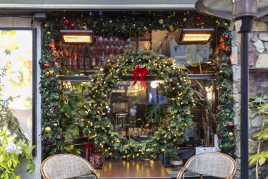 London, UK - November 23, 2024. The window display of a cafe is adorned with a Christmas wreath made of fir branches, pinecones, and red berries. Outdoor seating features cafe tables and gas lamp heaters. clipart