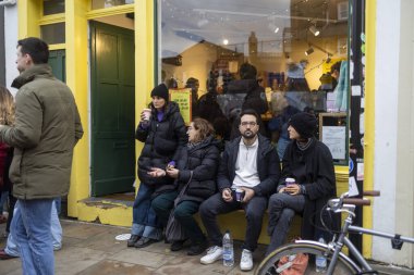 Londra, İngiltere - 28 Kasım 2024, Covent Garden Market 'te sarı duvarı olan bir dükkanın yanındaki bankta oturan insanlar.