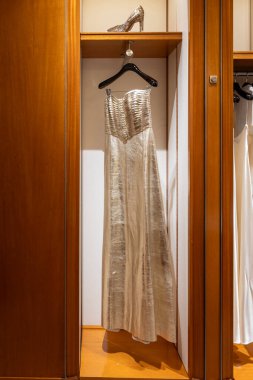 Sırtı açık ve zemin hizasında dizayn edilmiş gümüş bir gece elbisesi ahşap bir gardıropta asılı, dışarı çıkmaya hazır..