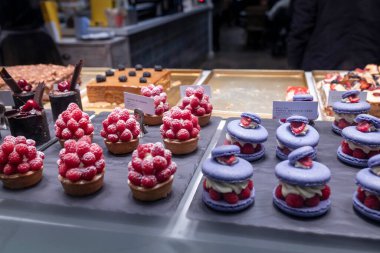 Londra, İngiltere - 1 Aralık 2024, fırınlanmış mallarla dolu bir aşçılık kafesi. Pudra şekerli lezzetli ahududu tartları kremayla dolu mor badem kurabiyeleri ve çikolatalı pastalar.