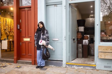 Londra, İngiltere, 1 Aralık 2024, Kareli yün ceketli genç bir Asyalı kadın Columbia Çiçek Pazarı 'nın renkli bir duvarının yanında poz veriyor..