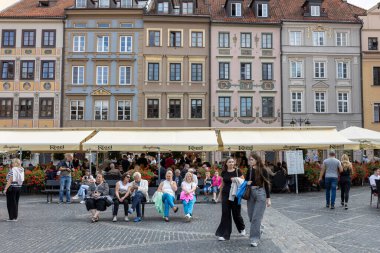 Varşova, Polonya - 2 Eylül 2024, Turistler Eski Şehir meydanında bir bankta oturuyorlar. İnsanlar restoranlarda yemek yiyor. Büyük bir karga.