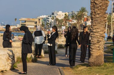 Siyah pantolon ve beyaz gömlek giymiş bir grup Arap kadın deniz kenarında selfie çekiyor..