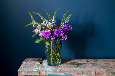 Veronica ve Ay tozu karanfillerinin düğün buketi kaba ahşap bir bankta, koyu mavi bir arka planda cam bir vazoda. Metin için boşluk
