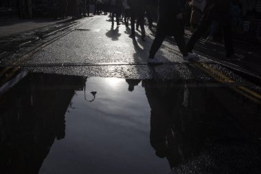 Tanınmayan insanlar büyük bir su birikintisinden kaçarak yol boyunca ilerliyorlar ve yansımaları asfalttaki su birikintisinde görülebiliyor. Şehirde iş çıkış saati