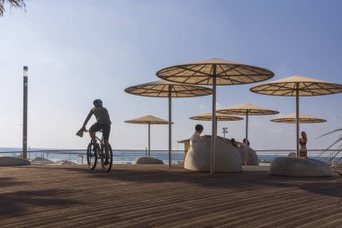 Tel Aviv, İsrail, 20 Kasım 2024, insanlar plajdaki dinlenme odasındaki büyük koltuklarda rahatlıyorlar. Bisiklet süren bir adam. Arka planda siluetler.