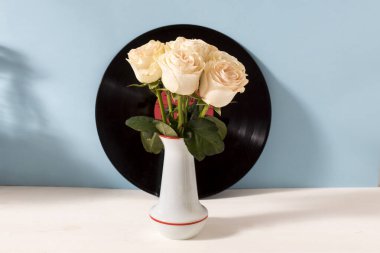 Beyaz seramik vazoda bir buket pembe gül mavi arka planda siyah plak plaklı ve beyaz masalı. Mesaj için yer var. Gelin buketi..