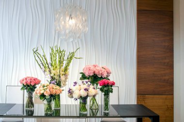 Modern bir iç mekanda cam bir yemek masasında güller, ortancalar ve gladioluslarla zarif bir çiçek aranjmanı.