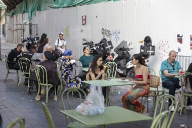 Kudüs, İsrail, 16 Mart 2025, Purim şehirde. Old City 'deki sokak kahvesi. Erkekler ve kadınlar demir masalarda oturup kola içerler ve bir motosikletin yanındaki inşaat duvarına karşı öğle yemeği yerler..