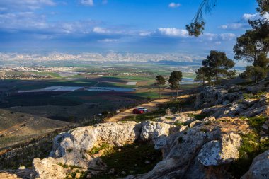 Gilboa Dağı 'ndan, çamların, bitkilerin, vadinin ve Ürdün dağlarının manzaralı manzarası, gökyüzünde bulutlar ve aşağıdaki tarım arazileri.
