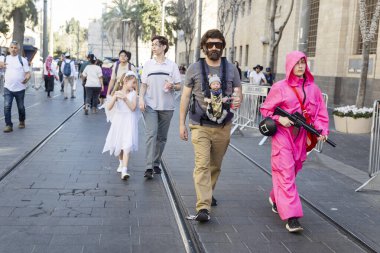 Kudüs, İsrail 16 Mart 2025. Şehirde Purim. Bir adam askıda bir bebek taşıyor. Pembe tulumlu ve tüfekli kız arkadaşı şehir sokaklarında.