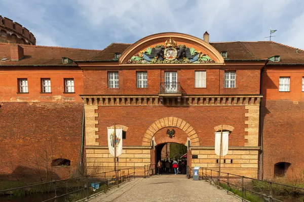 Berlin, Almanya - 30 Mart 2024: Ortaçağ Fuarı: Easter Knight Spectaculum. Spandau Kalesi 'nde bir ortaçağ pazarı. Turistler köprüden geçerek Spandau Kalesi 'nin girişine doğru yürüyorlar. Arka planda kırmızı tuğla duvarlar ve kule var..