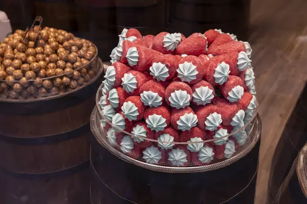 Yakın plan çekimde, içi şekerle dolu iki cam tabak görünüyor. Koyu tahta fıçıların üzerinde sergileniyor. Tabak kırmızı çilek şeklinde şekerler içeriyor, her birinin üzerinde küçük, beyaz bir girdap var.. 