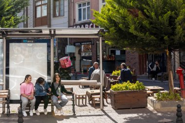 İstanbul, Türkiye, 2 Mayıs 2024, Balat ilçesinde sokak sahnesi, otobüs durağında oturan gençlerin, bir kafenin açık masalarında oturan erkeklerin ve pembe sırt çantalı bir teslimatçının, güneşli bir günde.