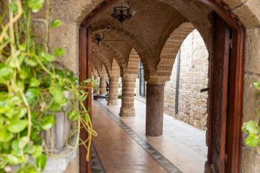 Tel Aviv-Jaffa, İsrail. 9 Nisan 2025, Al bahar Camii. Sütunları, desenli döşemeleri ve asılı lambaları olan kemerli taş bir koridor, eski jaffa yemyeşil bitkilerle dolu açık bir kapı aralığından görülüyor., 