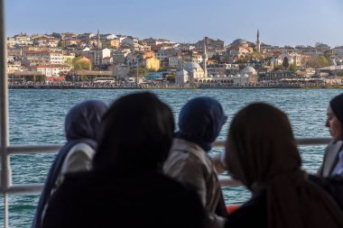 İstanbul, Türkiye, 5 Nisan 2024, İstanbul, Türkiye 'de bir feribota binen yolcular, kıyısı boyunca camileri ve binaları bulunan şehir manzarasına bakıyorlar. Bazıları başörtüsü takıyor.