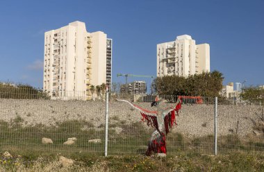 Hayfa, İsrail, 16 Nisan 2025, bir kuşa benzeyen büyük, renkli, iplik bombalı sanat kurulumu, arka planda apartman binaları bulunan kentsel ya da banliyö bölgesindeki bir tel örgüye bağlıdır..