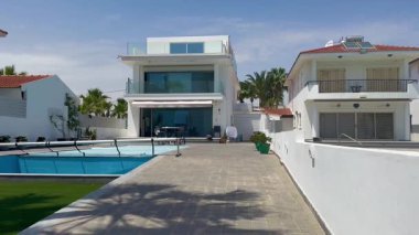 Larnaca, Kıbrıs, 8 Mayıs 2025. Önünde balkonları ve yüzme havuzu olan iki katlı beyaz villalar. Bir palmiye ağacı rüzgarda sallanan bir gölge bırakır..
