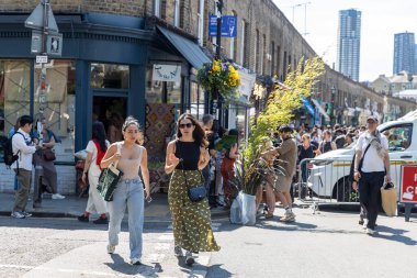 Londra, İngiltere, 10 Mayıs 2025. Güneşli bir günde, Londra, Shoreditch 'te iki şık kadın alışveriş ve market tezgahlarıyla dolu bir caddede yürüyor..