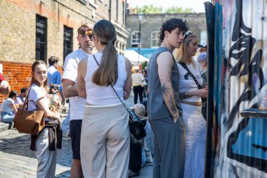 Londra, İngiltere, 10 Mayıs 2025 'te, çeşitli bir grup insan güneşli bir günde Londra' daki Columbia Road Çiçek Pazarı 'ndaki graffiti sanatının yanında canlı bir sokakta toplandı..