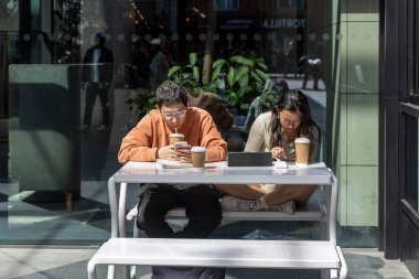 Londra, İngiltere, 10 Mayıs 2025, iki genç yetişkin küçük bir açık hava masasında oturuyor, her biri elektronik cihazlarına bakıyor ve Londra 'daki kahve fincanlarından içiyor..
