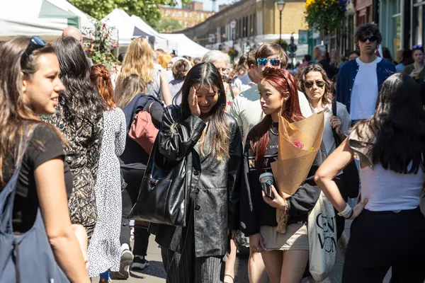 Londra, İngiltere, 5 Mayıs 2025, İnsanlar kalabalık bir sokak pazarından geçiyor. Asyalı genç kadın birbiriyle konuşuyor.
