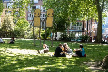 Londra, İngiltere, 5 Mayıs 2025, İnsanlar parktaki Stik heykellerinin yakınındaki çimlerin üzerinde dinleniyor. Stik, Shoreditch 'teki Hoxton Meydanı' nda bir heykel yaptı..