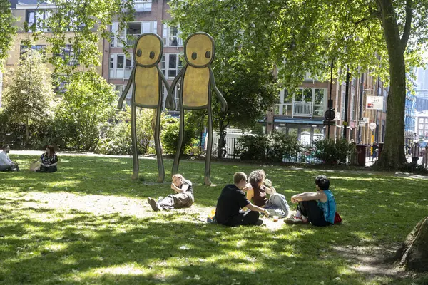 Londra, İngiltere, 5 Mayıs 2025, İnsanlar parktaki Stik heykellerinin yakınındaki çimlerin üzerinde dinleniyor. Stik, Shoreditch 'teki Hoxton Meydanı' nda bir heykel yaptı..