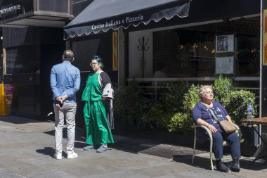 Londra, İngiltere, 5 Mayıs 2025, yeşil ipek takım elbiseli şık bir kadın bir erkekle konuşuyor. Yaşlı bir kadın bir İtalyan kafesinin önündeki masada güneşleniyor..