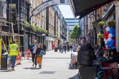 Londra, İngiltere, 5 Mayıs 2025, Carnaby Caddesi 'ndeki Alışveriş ve Turistler. Parlak giyimli bir adam ve kadın inşaat işçilerinin yanından geçerek bir evi yeniliyorlar.