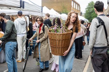 Londra, İngiltere, 7 Mayıs 2025, uzun dalgalı saçlı genç bir kadın, Columbia Çiçek Yolu Pazarı 'ndan alınmış bir sepet çiçek taşıyor.