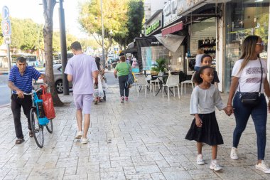 Petah Tikva, İsrail, 12 Haziran 2025, İsrail 'de Şehir Kaldırımında Yürüyenler. Mavi tişörtlü yaşlı bir adam bisiklet itiyor. Çocuklarıyla birlikte Etiyopyalı bir anne
