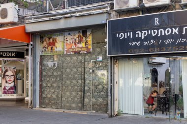 Petah Tikva, İsrail, 12 Haziran 2025, Dükkan Cephesi ve Çocuk. Kırmızı tişörtlü bir çocuk kozmetoloji kliniğinde annesini bekliyor.