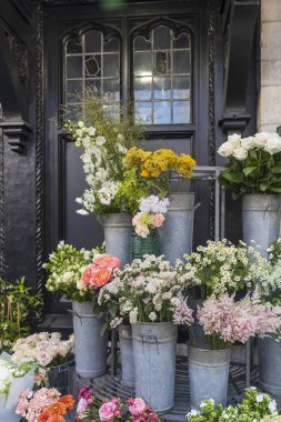 Renkli çiçeklerden ve yeşilliklerden oluşan geniş bir çeşitlilik, arka planda dekoratif bir pencere bulunan bir çiçekçi dükkanının dışındaki galvanize metal kovalarda düzenlenir..