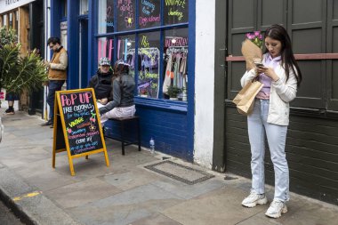 Londra, İngiltere, 9 Mayıs 2025 Genç Asyalı bir kadın bir dükkanın önünde telefonuna bakıyor..