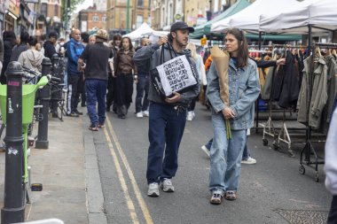Londra, İngiltere, 9 Mayıs 2025 Bustling Outdoor Market 'te bir çift yürüyor. Bir genç erkek ve bir genç yetişkin kadın, her ikisi de Kafkasyalı, görünür. Arka planda diğer yetişkinler var..