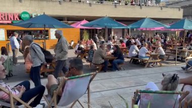 Londra, İngiltere, 7 Mayıs 2025, sıcak bir Mayıs günü. İnsanlar Ulusal Tiyatro 'nun yakınındaki bir kafede iskemlede oturuyorlar. Sohbet ederler, yerler ve şarap içerler..