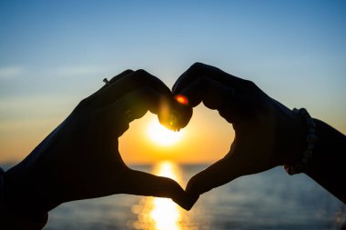 Siluetli eller, okyanusun üzerinde altın bir günbatımına karşı bir kalp şekli oluşturuyor. Güneş ışığı, sakin suya yansıyor..