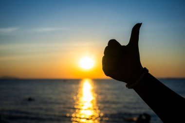 Silüetli el, okyanusun üzerinde canlı bir günbatımına karşı baş parmak işareti yapıyor. Altın güneş ışığı suya yansıyor..