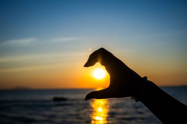 Silüetli el, okyanusun üzerinde parlayan bir günbatımının önünde, altın güneş ışığını yansıtan yarım kalp jesti oluşturuyor.