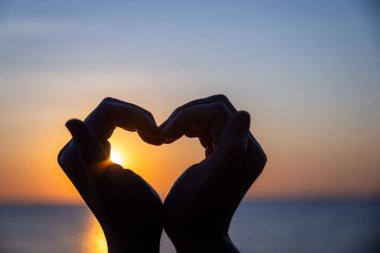 Siluetli eller bir kalp şekli oluşturuyor. Okyanusun üzerinde batarken parlayan güneşi mükemmel bir şekilde çerçeveliyor. Huzurlu bir arkaplan renkleri ve durgun sularla..