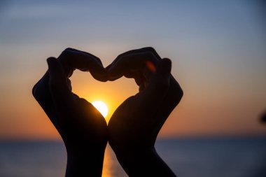 Siluetli eller bir kalp şekli oluşturuyor. Okyanusun üzerinde batarken parlayan güneşi mükemmel bir şekilde çerçeveliyor. Huzurlu bir arkaplan renkleri ve durgun sularla..