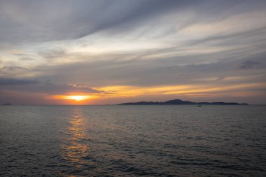 Güneş ufkun altına batıyor, sakin denizin üzerine altın bir parıltı saçıyor. Yalnız bir yelkenli körfez boyunca huzur içinde süzülürken hafif bir esinti suyu dalgalandırır. Martılar tepeden ağlar, sakin atmosferi artırır..