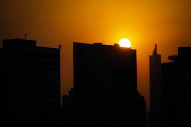 Canlı turuncu bir günbatımına karşı şehrin silueti. Yüksek binalar parlak gökyüzüne karşı dizilmiş dramatik ve büyüleyici bir sahne arkaplanı oluşturuyorlar.