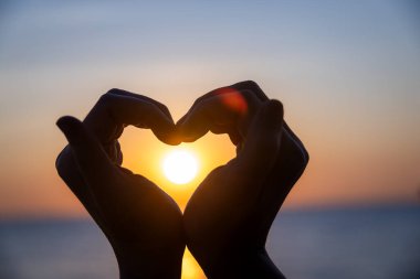Siluetli eller bir kalp şekli oluşturuyor. Okyanusun üzerinde batarken parlayan güneşi mükemmel bir şekilde çerçeveliyor. Huzurlu bir arkaplan renkleri ve durgun sularla..
