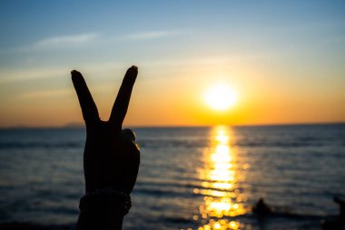 Siluetli el, okyanusun üzerinde parlayan bir günbatımına karşı barış işareti yapıyor. Altın ışık suya yansıyor.