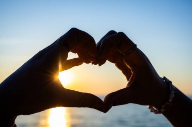 Siluetli eller, okyanusun üzerinde altın bir günbatımına karşı bir kalp şekli oluşturuyor. Güneş ışığı, sakin suya yansıyor..